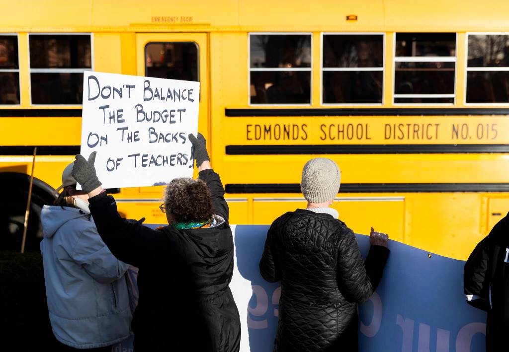 Melody Schneider holds a sign protesting pay cuts to teachers as an Edmonds School District bus passes by during an Edmonds College faculty union rally as part of a national day of action outside of the Lynnwood Event Center on Tuesday, March 4, 2025, in Lynnwood, Washington. (Olivia Vanni / The Herald)